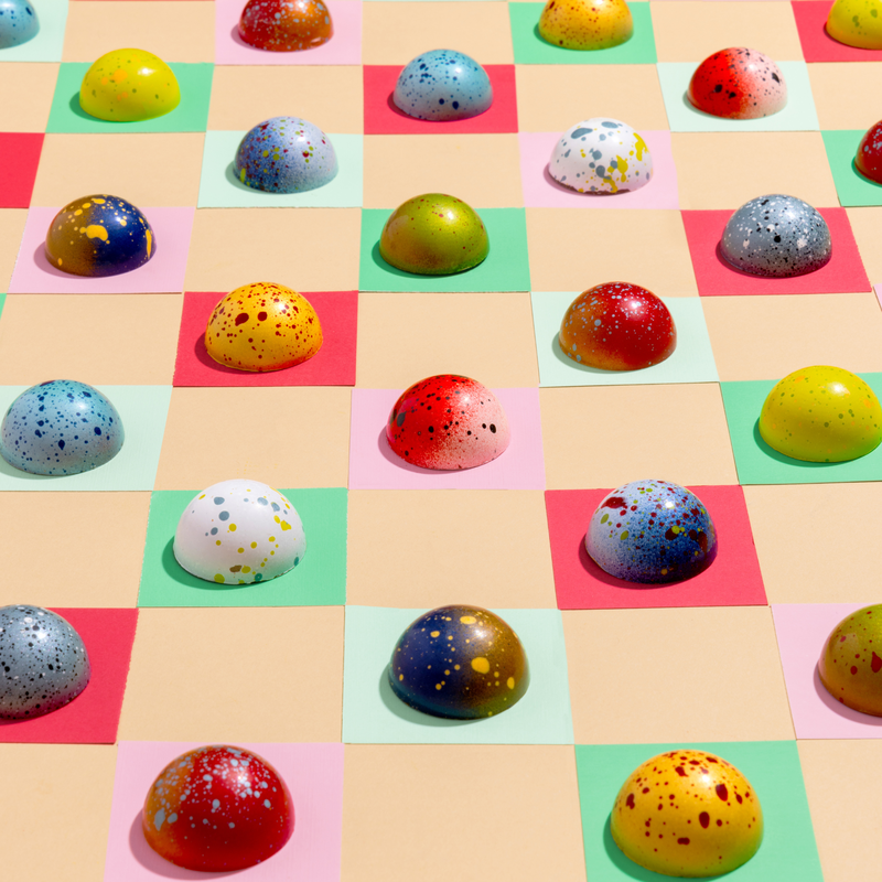 Individual chocolates from the Holiday Gift Box displayed on a checkerboard background. Each chocolate is lovingly designed to resemble Christmas ornaments and evoke Yuletide nostalgia.